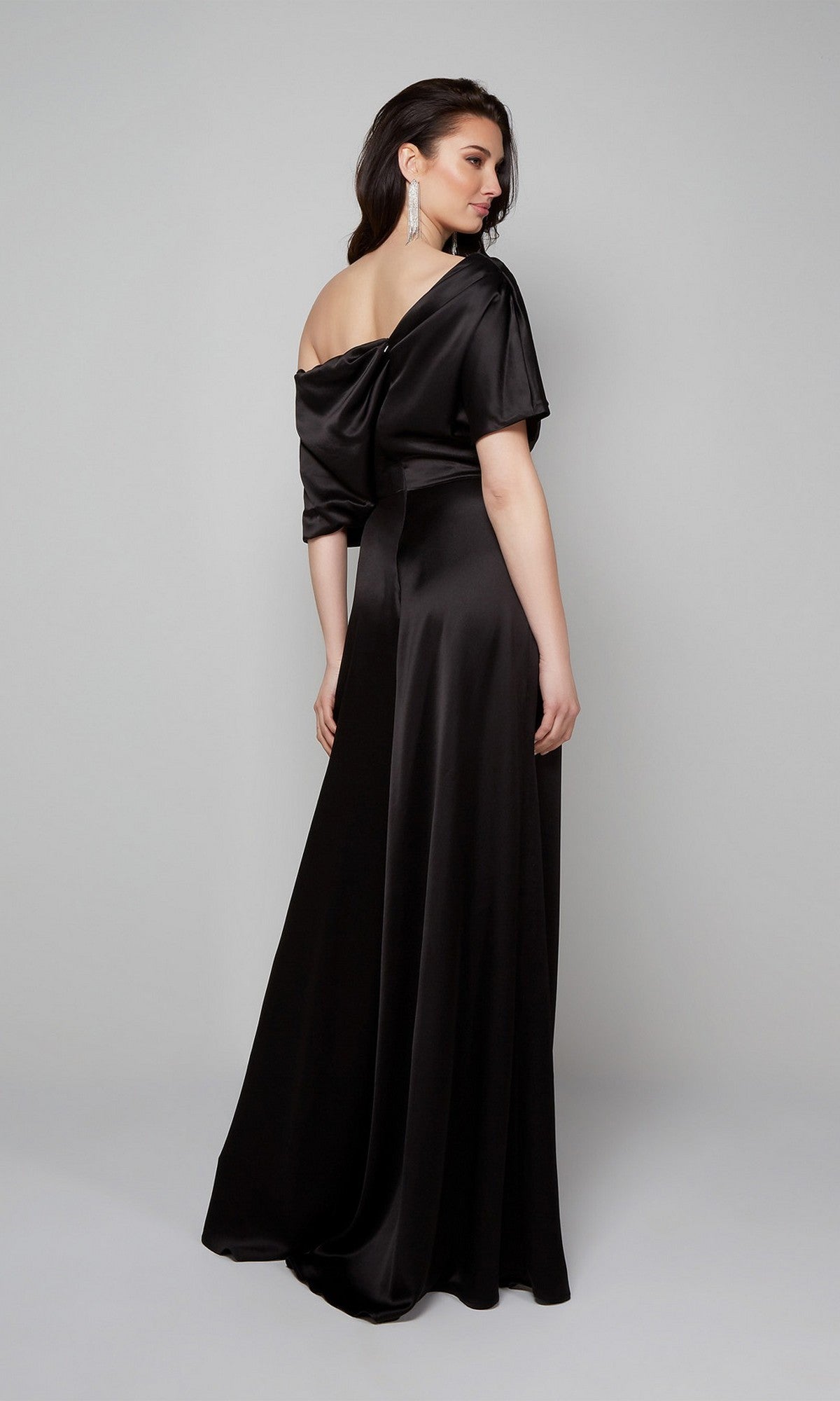 A woman stands in a studio, facing away from the camera, wearing the Alyce 70019 long formal jumpsuit with off-the-shoulder black satin, her hair down and accessorized with long earrings.